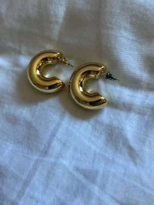 Chunky guldfärgade hoops örhängen - Säljer ett par chunky hoops örhängen i guldfärgad metall. De har en rundad, tjock form och stängs med klassisk stift. Perfekta för dig som gillar statement-smycken och vill addera lite extra bling till din look.