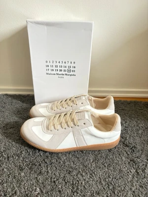 Maison Margiela vita sneakers - Stilrena sneakers från Maison Margiela i vitt och beige med ovandel i läder och mocka. Klassisk låg modell med beige snörning och gummisula. Perfekta för dig som gillar minimalistisk och clean stil.