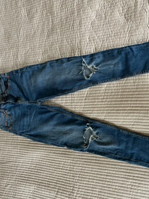 Blå slitna jeans med hål XS - Säljer ett par blå jeans i storlek XS från 157 med snygga slitningar och stora hål på båda benen. Jeansen är gjorda i mjukt denimtyg. Perfekta för en avslappnad och trendig look.