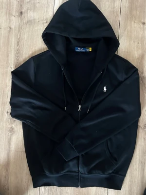 Svart hoodie från Polo Ralph Lauren - Säljer en svart hoodie från Polo Ralph Lauren med dragkedja framtill och klassisk huva. Tröjan har det ikoniska vita Polo-logotrycket på bröstet och ribbade muddar vid ärmar och nederkant. Perfekt för en avslappnad och stilren look.