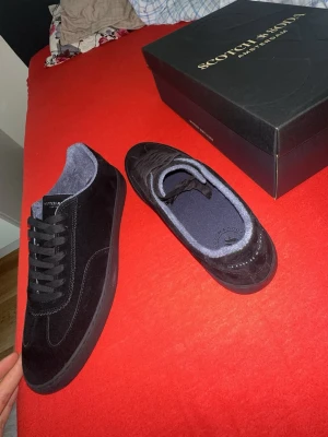 Svarta Scotch & Soda sneakers i mocka - Stilrena svarta sneakers från Scotch & Soda i mjuk mocka med svart sula och svarta skosnören. Insidan är fodrad i mörkgrått tyg för extra komfort. Klassisk låg modell som passar till det mesta och ger en clean look.