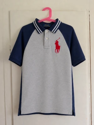Polo Ralph Lauren piké – strl M (10–12 år) - Snygg Polo Ralph Lauren-piké i grått och marinblått med klassisk krage och stor röd broderad hästlogga framtill. På ryggen finns ett stort vitt “Polo RL P 1930 Athletics”-tryck. Storlek: M / 10–12 år Färg: Grå & marinblå Märke: Polo Ralph Lauren Skick: Fint begagnat skick utan defekter! Sportig och klassisk modell som passar perfekt till jeans eller shorts. 💙❤️ Skriv gärna vid frågor eller om du vill ha fler bilder/mått!