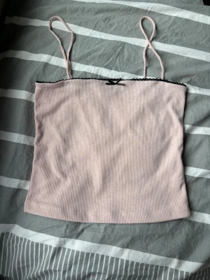 Ljusrosa ribbat linne - Söt ljusrosa ribbad topp med tunna spaghettiband och svart spetskant upptill. Dekorerad med en liten svart rosett framtill. Perfekt för sommaren och enkel att matcha med olika stilar.