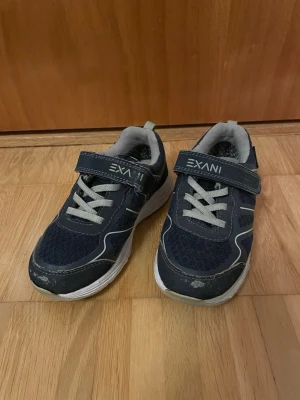 Mörkblå sneakers från Exani - Snygga mörkblå sneakers från Exani med vita detaljer och grå meshpartier. Skorna har kardborreband och elastiska skosnören för enkel påtagning. Yttersulan är vit och ger en sportig look. Perfekta för dig som vill ha bekväma och stilrena sneakers till vardags.