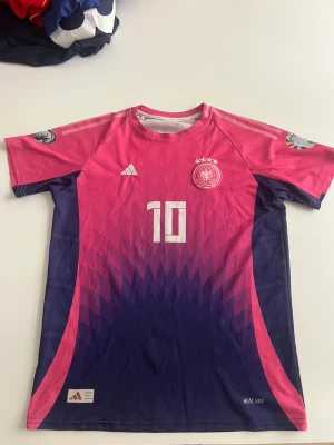 Tysklands 2024 borta musiala - Snygg Tyskland fotbollströja från Adidas i rosa och lila med nummer 10 och Musiala på ryggen. Tröjan har korta ärmar, rund hals och tryckta detaljer som landslagslogga och stjärnor. Tillverkad i lätt och ventilerande material, perfekt för träning eller match.