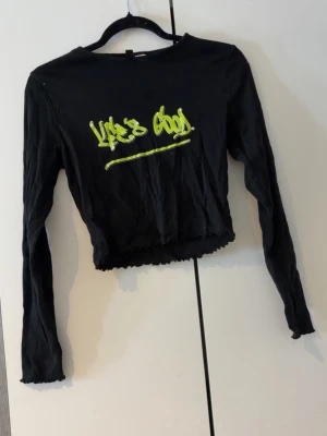 Svart croppad långärmad topp Divided - Svart croppad långärmad topp från Divided med limegrönt tryck 'Life's Good' framtill. Toppen har en tight passform och vågig kant nertill för en trendig look. Perfekt till jeans eller kjol för en cool streetstyle.