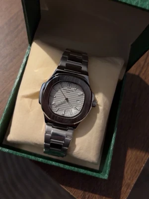 Klocka - Stilren klocka i silver med länkarmband och rundad fyrkantig boett. Urtavlan är ljus med diskreta linjer och tydliga indexmarkeringar. Perfekt accessoar för dig som gillar en klassisk och modern look.