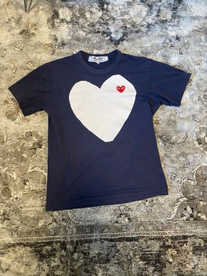 Mörkblå t-shirt med hjärta - Mörkblå t-shirt med stort vitt hjärta på bröstet och en liten röd hjärtdetalj. Klassisk rund hals och korta ärmar. Perfekt för dig som gillar grafiska tryck och vill sticka ut lite extra.