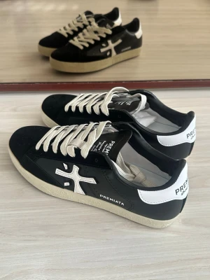 Svarta Premiata sneakers med vit sula - Snygga svarta sneakers från Premiata med vit sula och vita detaljer. Skorna har en låg profil, vita skosnören och en tydlig logga på sidan. Materialet är en mix av läder och mocka för en stilren look.