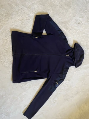 Mörkblå hoodie från Prada - Snygg mörkblå hoodie från Prada med dragkedja framtill och huva. Ärmen har en cool ficka med dragkedja och den klassiska Prada-loggan i svart och silver. Materialet ser ut att vara en mix av bomull och nylon, vilket ger en sportig och exklusiv känsla.