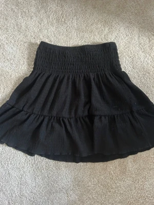 Svart volangkjol med resårmidja - Kort svart kjol med bred smockresår i midjan och volangdetaljer. Kjolen har en luftig och flowy passform som ger en avslappnad men ändå trendig look. Perfekt att matcha med en crop top eller stickad tröja.