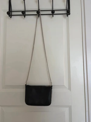 Svart kuvertväska med kedja - Stilren svart kuvertväska i slätt material med silverfärgad kedjerem och dekorativa silverkulor längs kanterna. Perfekt för att lyfta din outfit med en edgy touch. Väskan har en rektangulär form och minimalistisk design.