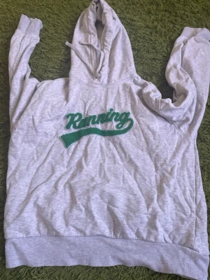 Grå hoodie med grön text Running - Säljer en ljusgrå hoodie med huva och dragsnören. På bröstet står det 'Running' i grön text med ett sportigt tryck. Hoodien har ribbade muddar vid ärmslut och nederkant. Perfekt för chill dagar eller när du vill ha en avslappnad stil.