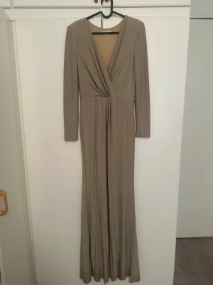 Glittrig långklänning med v-ringning och slit i sidan av benen - Snygg långklänning i glittrigt beige tyg med djup v-ringning och långa ärmar. Klänningen har omlott-detalj framtill och ett elegant fall. Perfekt för dig som vill sticka ut på festen.