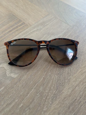 Ray-Ban Erika - Snygga solglasögon från Ray-Ban med leopardmönstrad båge i bruna och svarta toner. Glasen är runda och har en mörk toning som ger en cool vibe. Tunn metallbåge på sidorna för en modern look. Perfekta för soliga dagar!