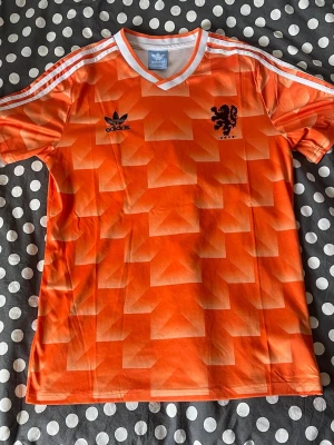 Orange Adidas Holland fotbollströja - Säljer en ikonisk orange fotbollströja från Adidas med Nederländernas lejonmärke på bröstet. Tröjan har ett retroinspirerat geometriskt mönster, vita ränder på axlarna och V-ringad krage. Perfekt för dig som älskar klassiska landslagströjor.