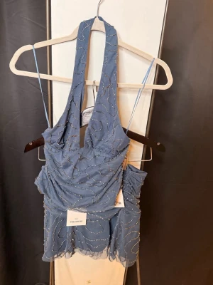 Blå glittrigt set - Supersnygg blå croppad linnetopp och kjol med glittriga detaljer och halterneck. Tunn mesh med vågigt mönster och transparent känsla. Perfekt för dig som vill sticka ut lite extra. Toppen är strl XS och kjolen S. Aldrig använt !