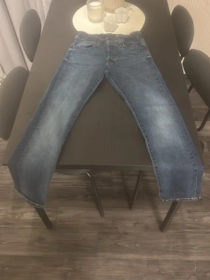 Blå Jack and Jones jeans - Säljer ett par blå jack and Jones jeans med rak passform och klassisk femficksdesign. Jeansen har en något ljusare tvätt på låren och knäna för en snygg, avslappnad look. Tillverkade i slitstarkt denimtyg som passar perfekt till vardagsstilen.