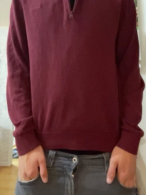 Hugo boss Mörkröd tröja med halv dragkedja - En stilren mörkröd långärmad tröja med halv dragkedja och hög krage. Tröjan har ribbade muddar vid ärmslut och nederkant. Perfekt för lager-på-lager och enkel att matcha med jeans eller chinos.