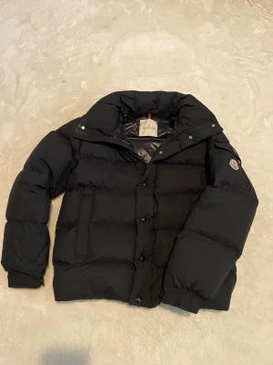 Svart dunjacka från Moncler - Snygg svart dunjacka från Moncler med hög krage, dragkedja och tryckknappar framtill. Jackan har två sidofickor, ribbade muddar och klassisk Moncler-logga på ärmen. Perfekt för kalla vinterdagar tack vare det tjocka dunfodret.