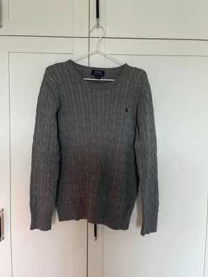 Grå kabelstickad tröja Polo Ralph Lauren - Snygg grå kabelstickad tröja från Polo Ralph Lauren med rund halsringning och klassisk marinblå logga broderad på bröstet. Tröjan har ribbade muddar vid ärmslut och nederkant. Perfekt för lager på lager och stilrena outfits.