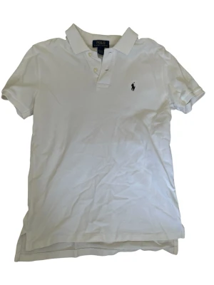 Vit pikétröja från Polo Ralph Lauren - Klassisk vit pikétröja från Polo Ralph Lauren med korta ärmar, krage och knappar framtill. Tröjan har den ikoniska broderade loggan på bröstet och är gjord i mjukt bomullsmaterial. Perfekt till jeans eller shorts för en clean look. Den är i använt skick men väl omhändertagen. Storleken är 10-12 år men jag skulle säga att den passar om man är ungefär 160-170 cm lång. Pris kan diskuteras!