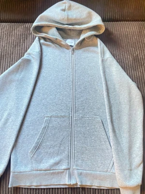 Grå zip hoodie från Jack & Jones - En stilren grå hoodie från Jack & Jones med dragkedja framtill och två stora fickor. Klassisk huva och mjukt material som känns skönt mot huden. Perfekt för en avslappnad look och enkel att matcha med jeans eller joggers.