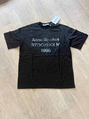 Svart Acne Studios t-shirt - Säljer en svart t-shirt från Acne Studios med tryck 'Acne Studios STOCKHOLM 1996' i vitt på bröstet. Klassisk passform med korta ärmar och rund hals. Perfekt för dig som gillar stilrena och trendiga plagg.