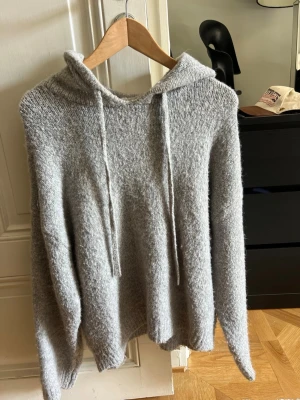 Grå stickad hoodie med luva - Mysig grå stickad hoodie med luva och dragsko. Tröjan har lång ärm och är gjord i ett mjukt, fluffigt material som känns skönt mot huden. Perfekt för kyliga dagar när du vill vara både bekväm och snygg.