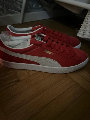 Röda Puma Suede sneakers - Snygga röda Puma Suede sneakers med vit sula och klassisk vit formstripe på sidan. Ovandelen är i mocka och skorna har röda skosnören samt guldfärgad Puma-logga på sidan. Perfekta för en streetwear-look. Oanvända 