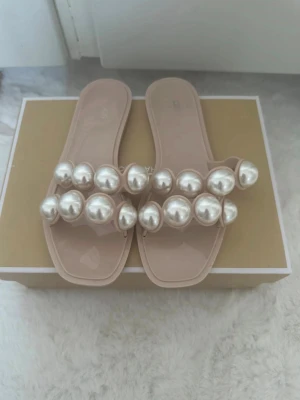 Beige sandaler med pärldekor - Säljer ett par trendiga beige sandaler med två remmar dekorerade med stora vita pärlor. Sandalerna har fyrkantig tå och är tillverkade i ett glansigt syntetmaterial. Perfekta för dig som vill sticka ut med en unik och snygg detalj på fötterna.