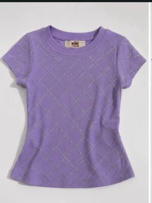Lila ribbad t-shirt med pärlor - Snygg lila t-shirt med korta ärmar och ribbad struktur. Framsidan har ett rutmönster dekorerat med små pärlor som ger en extra touch. T-shirten har en rund halsringning och figurnära passform, perfekt för en trendig vardagslook.