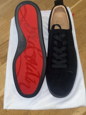 Svarta sneakers med röd sula - Snygga svarta sneakers i mocka från Christian Louboutin med den ikoniska röda sulan och svarta skosnören. Låg modell med stilren design och diskreta detaljer. Perfekta för dig som vill ha något exklusivt och trendigt.