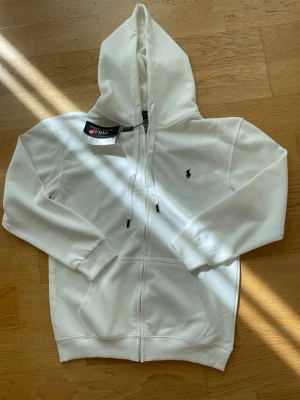 Vit hoodie med dragkedja och huva - Snygg vit hoodie med dragkedja och huva. Klassisk design med två fickor framtill och justerbara snören i huvan. Liten broderad logga på bröstet. Tillverkad i mjukt material som känns skönt mot huden. Perfekt för en avslappnad och stilren look.