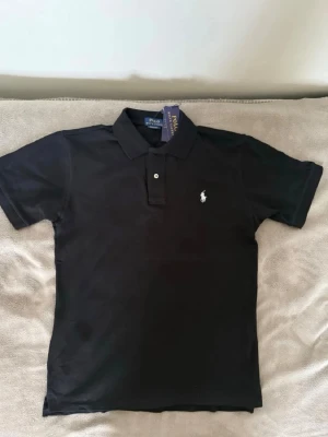 Svart pikétröja från Ralph Lauren - Klassisk svart pikétröja från Ralph Lauren med vit broderad logga på bröstet. Tröjan har krage, två knappar framtill och korta ärmar. Perfekt till jeans eller shorts för en clean och stilren look.