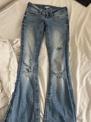 Ljusblå lowwaist jeans med hål och vingar på fickan - Säljer ett par ljusblå bootcut jeans med slitningar och hål på båda benen. Jeansen har klassisk femficksmodell, normalhög midja och utsvängda ben. Perfekta för en avslappnad och trendig look.