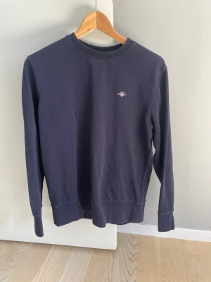 Mörkblå gant sweatshirt i fint skick - En stilren mörkblå sweatshirt med rund hals och liten broderad logga på bröstet. Tröjan har långa ärmar och ribbade muddar vid ärmslut och nederkant. Perfekt för en avslappnad och enkel look.