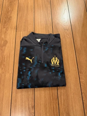 Svart träningströja Puma Marseille - Snygg svart träningströja från Puma med Olympique de Marseille-logga och guldfärgade detaljer. Tröjan har halv dragkedja, mönster i blått och svart samt hög krage. Perfekt för fotbollsträning eller sportiga dagar.