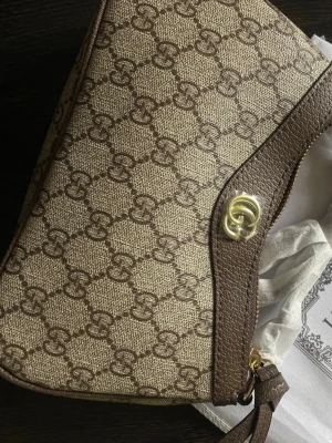 Beige och brun axelväska med GG-mönster - Snygg axelväska i beige och brunt med klassiskt GG-mönster och guldfärgad logga framtill. Väskan har detaljer i brunt läder och dragkedja upptill. Perfekt storlek för det viktigaste och en stilren accessoar till din outfit.