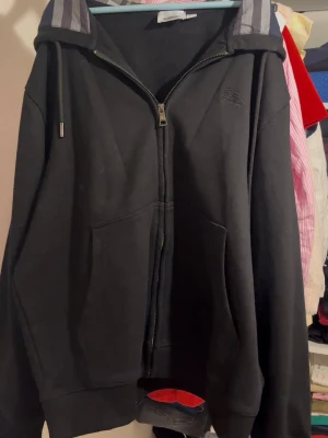 Burberry hoodie - Säljer en svart hoodie med dragkedja och kängurufickor. Huvan har ett snyggt rutmönstrat foder i blått, vitt och rött som ger en unik touch. Perfekt för dig som gillar klassisk stil med en twist. Materialet är mjukt och bekvämt, och passformen är avslappnad.
