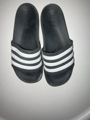 Svarta Adidas badtofflor med vita ränder - Klassiska svarta Adidas badtofflor med tre vita ränder över foten. Tillverkade i syntetmaterial med bekväm formad sula och öppen tå. Perfekta för stranden eller poolen och har den ikoniska sportiga looken.