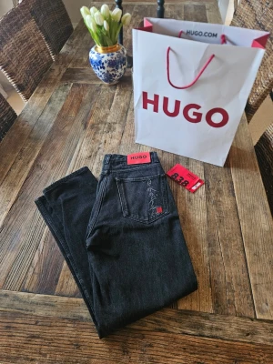 Svarta jeans från HUGO - Snygga svarta jeans från HUGO med raka ben och klassisk femficksdesign. Jeansen har en röd HUGO-label bak i midjan och en diskret japansk silvertext på bakfickan. Tillverkade i ett mjukt denimtyg som passar perfekt till streetwear eller en clean look. Köpta på MoS, väldigt snålt andvända ett nypris på 1799:- Hör av dig om du har några frågor!