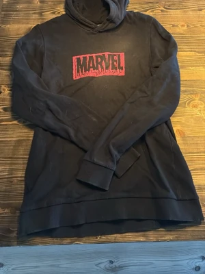 Svart Marvel hoodie med Thor-tryck - Säljer en svart hoodie med röd Marvel-logga på bröstet och en stor Thor-illustration på ryggen. Hoodien har huva och ribbade muddar vid ärmslut och nederkant. Perfekt för dig som gillar superhjältar och vill ha en bekväm och snygg tröja.