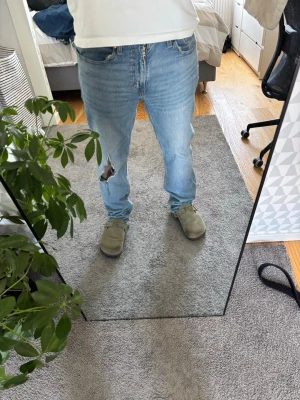 Ljusblå raka jeans med hål - Säljer ett par ljusblå jeans med rak passform och klassisk femficksdesign. Jeansen har ett stort hål på vänster lår och är tillverkade i denim. Perfekta för en avslappnad och trendig look.
