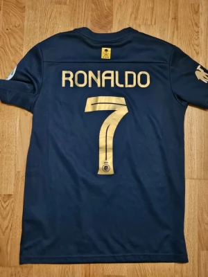 Mörkblå Al Nassr Ronaldo matchställ - Snyggt mörkblått matchställ från Al Nassr med guldiga detaljer. Tröjan har Ronaldo och nummer 7 på ryggen, samt klubbmärke och Nike-logga framtill. Setet inkluderar både tröja och shorts i lätt och ventilerande material, perfekt för fotbollsträning eller match.
