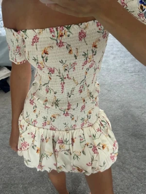 Offshoulder blommig kortklänning - Supersöt offshoulder kortklänning med smockad överdel och volangig kjol. Klänningen är vit med blommigt mönster i rosa, gult och grönt. Perfekt för varma dagar och har en romantisk vibe. Tillverkad i ett lätt och bekvämt material.