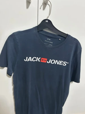 Mörkblå Jack & Jones t-shirt storlek S - Snygg mörkblå t-shirt från Jack & Jones med vit logga och röd detalj på bröstet. Klassisk rund halsringning och korta ärmar. Perfekt till jeans eller shorts för en avslappnad stil.
