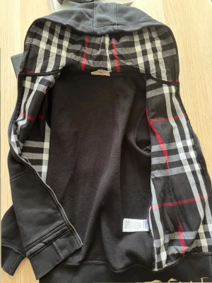 Svart hoodie med rutigt foder - Säljer en svart hoodie med dragkedja och huva. Insidan av huvan och öppningen har ett klassiskt rutigt mönster i svart, vitt och rött. Hoodien har två fickor framtill och ribbade muddar vid ärmar och nederkant. Perfekt för en chill och stilren look.