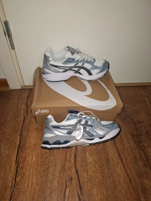 ASICS vita och grå sneakers - Snygga ASICS sneakers i vitt, grått och silver med svarta detaljer. Skorna har en sportig design med mesh och syntetmaterial, samt klassiska ASICS-ränder på sidorna. Sulan är vit med svarta och guldiga inslag undertill. Perfekta för en trendig streetstyle-look.