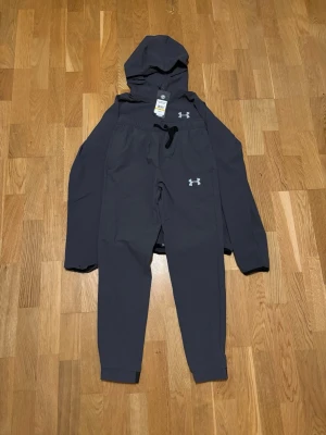 Mörkgrå träningsset från Under Armour - Snyggt mörkgrått träningsset från Under Armour med matchande hoodie och byxor. Hoodien har dragkedja och logga på bröstet, byxorna har resår i midjan och logga på benet. Perfekt för gymmet eller chill dagar.